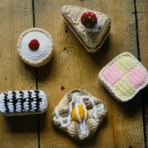 May include: A collection of crocheted food items, including a strawberry shortcake, a custard tart, a lemon meringue pie, a vanilla slice, and a jam and cream scone.