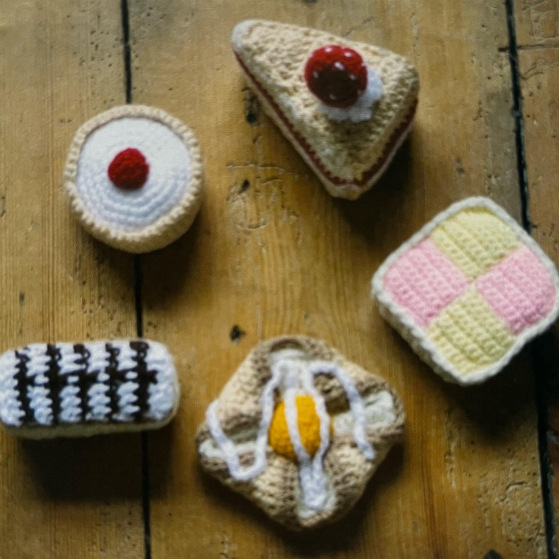 May include: A collection of crocheted food items, including a strawberry shortcake, a custard tart, a lemon meringue pie, a vanilla slice, and a jam and cream scone.