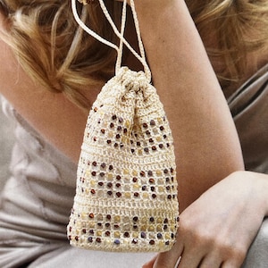 Könnte beinhalten: Eine kleine, gehäkelte Kordeltasche mit beigefarbener Basis und braunen, weißen und violetten Perlen, die in das Gewebe eingearbeitet sind.