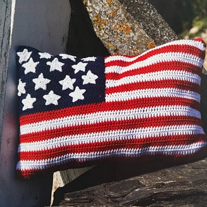 May include: A crocheted pillow in the shape of the American flag. The pillow is red, white, and blue with white stars on a blue field. The pillow is sitting on a wooden surface.