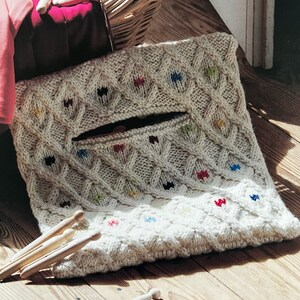 May include: A white knitted bag with a diamond pattern and colorful accents. The bag has a zippered pocket and is lying on a wooden surface.