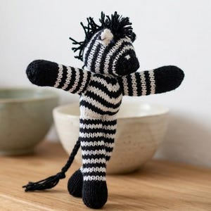 May include: A handmade knitted zebra toy in black and white stripes. The zebra has outstretched arms, a black mane, and a long tail. The toy is standing on a wooden surface, with a blurred background of bowls.