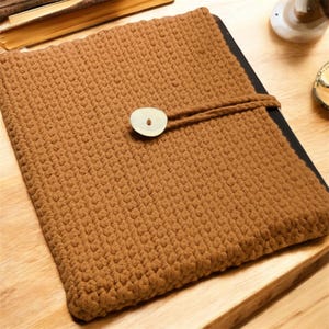 Puede incluir: Una funda de tableta marrón tejida a ganchillo con un cierre de botón de madera grande y redondo. La funda es rectangular y tiene una superficie texturizada. El botón está sujeto con un cordón marrón a juego. La funda está sobre una superficie de madera.