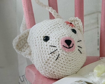 May include: A white crocheted cat toy with black eyes, a pink nose, and a red flower on its head. The toy is hanging from a pink wooden chair.
