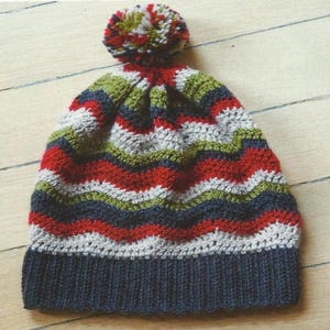 Puede incluir: Un gorro de punto a rayas con un pompón en la parte superior. El gorro está hecho en un patrón ondulado con rayas alternas de rojo, blanco, verde y azul.