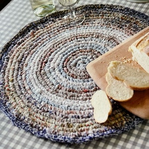 May include: A round, crocheted placemat with a blue, white, and brown color scheme. The placemat is made of recycled materials and has a unique, textured design. A wooden cutting board with sliced bread is on top of the placemat.