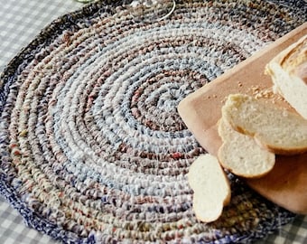 May include: A round, crocheted placemat with a blue, white, and brown color scheme. The placemat is made of recycled materials and has a unique, textured design. A wooden cutting board with sliced bread is on top of the placemat.