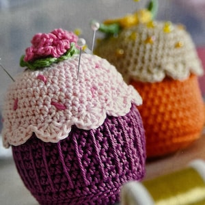 以下が含まれることがあります： 2つのクロシェで編まれたカップケーキの針山。1つはピンクとパープルで、上にピンクの花が飾られています。もう1つはオレンジとベージュで、黄色のビーズが付いています。