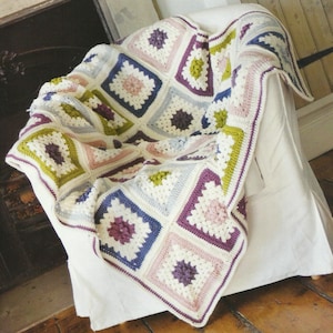 May include: A crocheted throw blanket with a granny square pattern in shades of green, purple, pink, and blue. The blanket is draped over a white armchair.