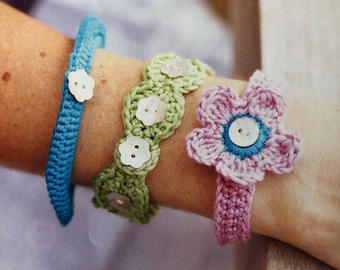 Puede incluir: Tres pulseras de ganchillo con detalles de botones. Las pulseras son en tonos de azul, verde y rosa. La pulsera rosa presenta un diseño de flor.