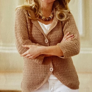 Peut inclure: Un blazer en crochet beige avec un motif texturé, doté d'un col cranté et de deux boutons. Le blazer a des manches trois-quarts et est associé à un haut blanc et un pantalon blanc. Un collier doré est visible.