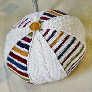 May include: A white crocheted pillow with a colorful striped pattern. The pillow is made with a variety of stitches, including a cable stitch and a bobble stitch.