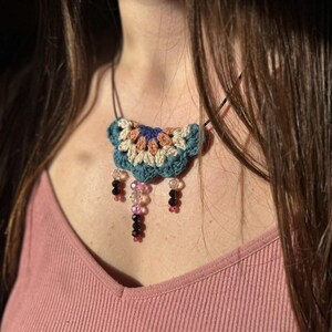May include: A handmade crochet necklace featuring a blue, beige, and orange floral design. The necklace has a black cord and dangling beads in shades of pink, clear, and black. The necklace is worn on a pink top.