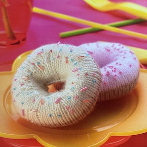 Op de afbeelding: Twee gebreide donuts, een beige met roze en blauwe sprinkles en een roze met roze sprinkles, op een geel bord.