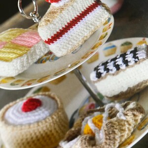 May include: A tiered cake stand with four crocheted cakes. The cakes are decorated with white icing, red cherries, and yellow and pink icing. The cakes are on a white cake stand with a gold trim.