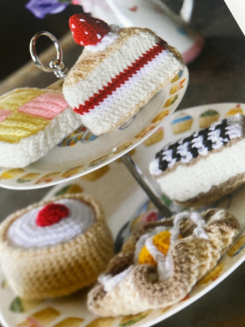 May include: A tiered cake stand with four crocheted cakes. The cakes are decorated with white icing, red cherries, and yellow and pink icing. The cakes are on a white cake stand with a gold trim.