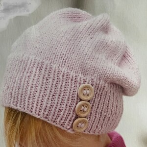 Op de afbeelding: Een lichtpaarse gebreide beanie met drie houten knopen aan de zijkant.