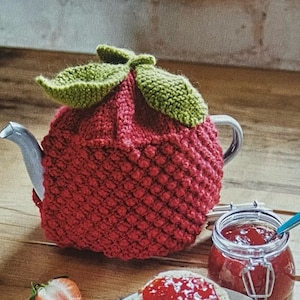 Puede incluir: Una funda de tetera de punto rojo con forma de fresa con hojas verdes. La funda está en una tetera con un pico blanco.