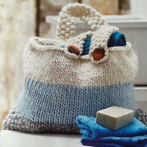 May include: A knitted tote bag with a white, blue and grey striped design. The bag has two brown buttons and a handle. The bag is filled with blue towels and a bar of soap.