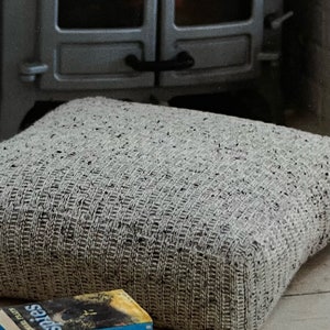 May include: A large, square, light gray knitted floor cushion with a textured pattern. A stack of books with blue and yellow covers is in front of the cushion. The background includes a fireplace.