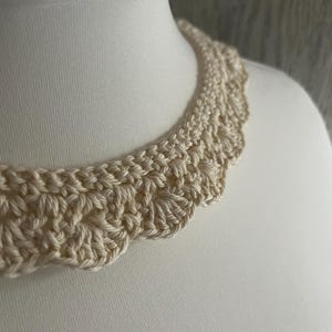 May include: A close-up of a crocheted collar in a cream color. The collar is made with a delicate stitch pattern and has a scalloped edge.