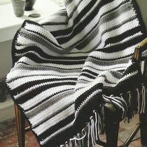 Op de afbeelding: Een gehaakte deken met zwarte, witte en grijze strepen, gedrapeerd over een houten stoel. De deken heeft een gefranjerde rand. Het handgemaakte item is een decoratief item voor in huis. De afmetingen van de deken zijn in centimeters.
