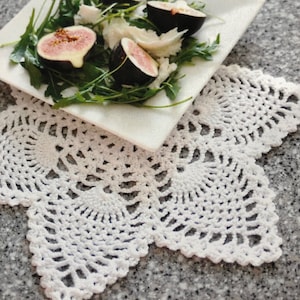 May include: A white crocheted doily with a scalloped edge sits on a table with a white plate of fresh salad with mozzarella cheese and figs.