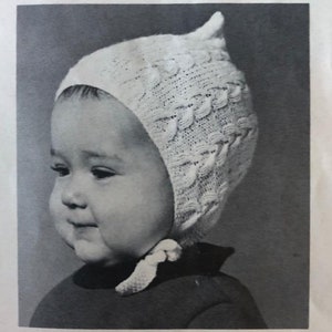 Op de afbeelding: Een zwart-witfoto van een baby die een witte gebreide muts met een puntige top draagt. De muts heeft een delicaat kabelbreipatroon.