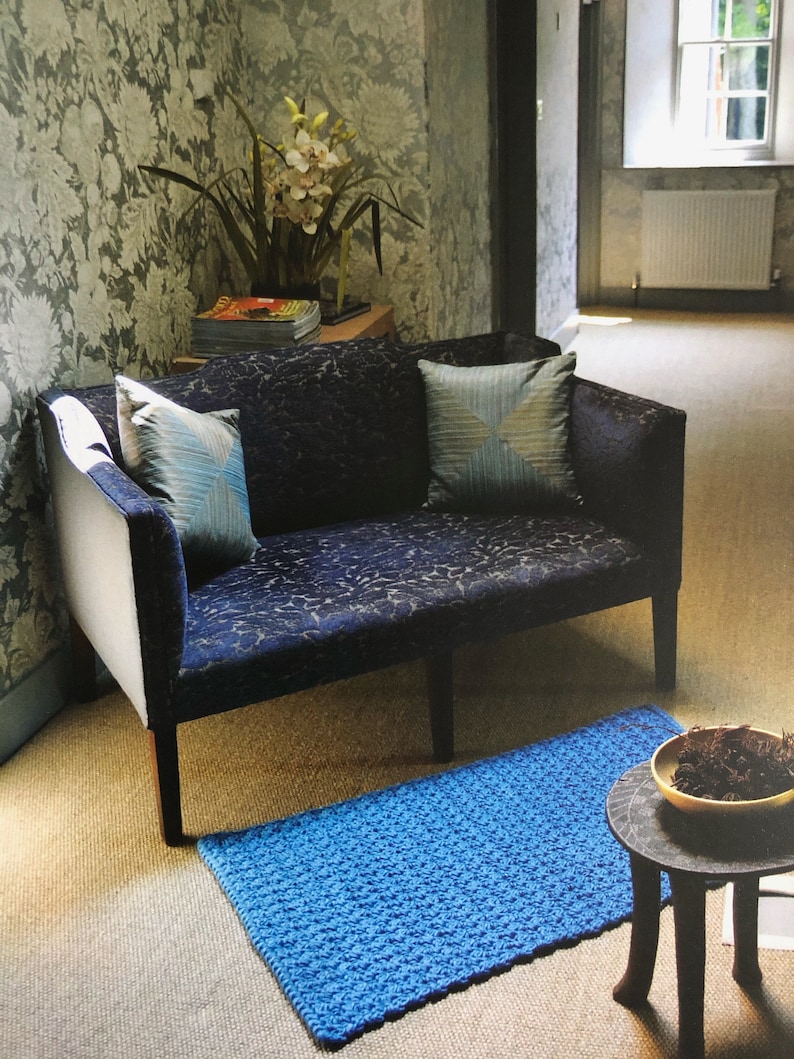 May include: A blue patterned sofa with two blue pillows sits in a room with a blue rug and a small wooden table with a bowl on top. The room has floral wallpaper and a window with a view of trees.