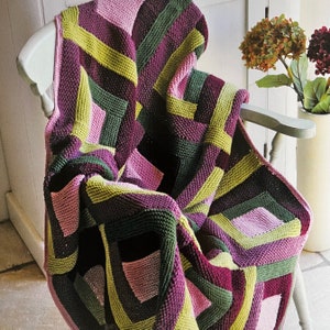 May include: A knitted throw blanket with a geometric pattern in shades of pink, green, and purple. The blanket is draped over a chair.