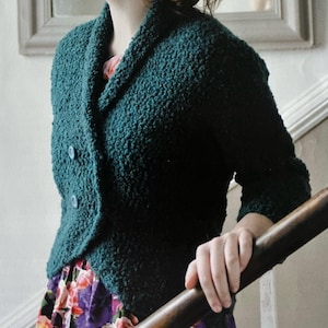 May include: A teal green textured knit jacket with a shawl collar and two light blue buttons. The jacket has three-quarter sleeves and is worn over a floral print dress. The image shows a close-up of the jacket.