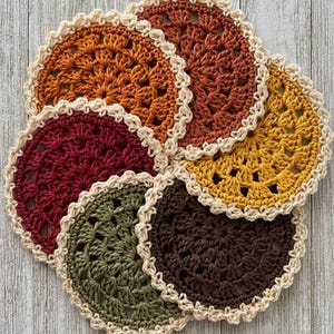 Könnte beinhalten: Ein Set aus sechs runden Häkeluntersetzern in herbstlichen Farben wie Rost, Orange, Senfgelb, Burgunderrot, Olivgrün und Dunkelbraun. Jeder Untersetzer hat ein zentrales Sternmuster und einen cremefarbenen, bogenförmigen Randabschluss.
