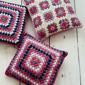 May include: Three crocheted square cushions in pink, white, and blue with a granny square pattern. The cushions are arranged on a white wooden surface.
