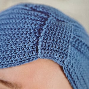 May include: Close-up of a blue crochet turban. The turban is made of a soft, textured yarn and features a ribbed design. The color is a muted blue, and the turban appears to be handmade. The style is a classic turban.
