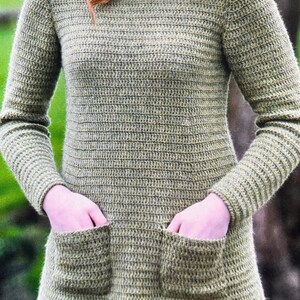 Könnte beinhalten: Ein grüner Strickpullover mit Rollkragen und zwei Taschen. Der Pullover ist aus weichem, strukturiertem Garn gefertigt.