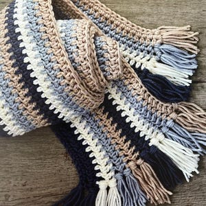 Op de afbeelding: Een handgehaakte sjaal in marineblauw, wit, lichtblauw en bruin. De sjaal heeft een gestreept patroon en franjes aan beide uiteinden. De zachte garen sjaal is ideaal om warm te blijven. De sjaal is ongeveer 180 cm lang.