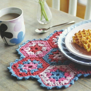 May include: A crocheted placemat in shades of pink, red, and blue with a scalloped edge. The placemat is set on a wooden table with a cup of coffee, a plate of waffles, and a vase of flowers.