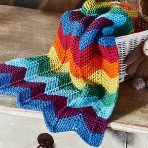 Pode incluir: Um cobertor de bebé de malha colorido com um padrão de chevron em tons de azul, verde, amarelo, laranja e vermelho.