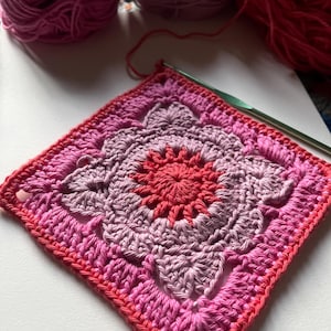 May include: A pink and red crochet square with a flower design in the center. The square is made with different shades of pink yarn and has a red center.