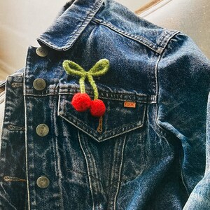 May include: A denim jacket with a green and red felt cherry pin on the pocket. The jacket has several metal buttons with the word "fcuk" on them.