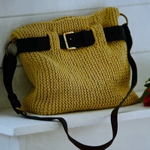 Könnte beinhalten: Eine senfgelbe Stricktasche mit einem schwarzen Lederriemen und einer Schnalle. Die Tasche hat einen langen Riemen zum Tragen über der Schulter.