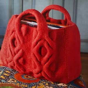 Pode incluir: Uma vibrante bolsa tote de feltro vermelho com duas alças, cheia de revistas. A bolsa apresenta um design geométrico em relevo na frente. A bolsa está sobre uma superfície estampada colorida.