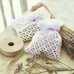 Op de afbeelding: Twee witte gehaakte lavendelzakjes met paarse strikken, een spoel gingham lint en gedroogde lavendeltakjes op een wit oppervlak.