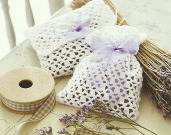 May include: Two white crocheted lavender sachets with purple ribbon bows, a spool of gingham ribbon, and dried lavender sprigs on a white surface.
