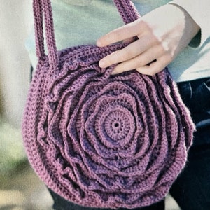 May include: A purple crocheted bag with a large flower design. The bag has a single strap and is made with a textured yarn.