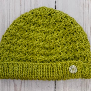 Puede incluir: Un gorro de punto de color verde con un borde doblado y un botón marrón.