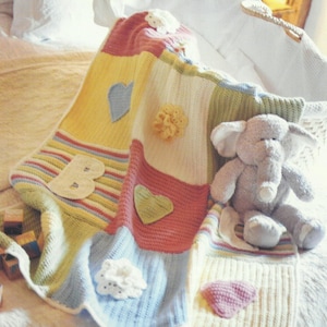 May include: A multi-colored crocheted baby blanket with a striped section, a heart, a flower, and the letter "B". A plush gray elephant toy is on top of the blanket.