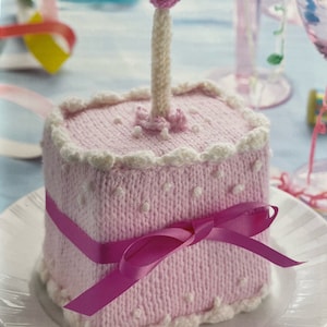 May include: A pink knitted cake with white frosting and pink polka dots. A pink ribbon is tied around the cake and a white knitted candle with a pink flame sits on top.