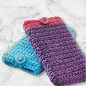 May include: Two handmade crochet phone cases. One is turquoise blue, the other is deep purple. Each case has a contrasting pink or blue button closure. The cases are made of soft yarn, ideal for protecting a mobile phone.