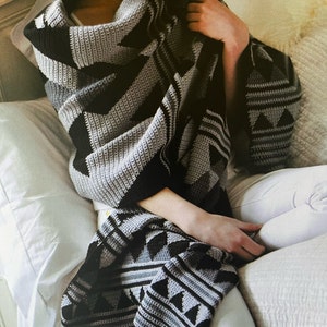 May include: A grey and black crocheted throw blanket with a geometric pattern. The blanket is draped over a person sitting on a bed.